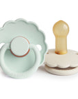 The FRIGG Daisy Natural Rubber Pacifier 2-Pack features two pastel, daisy-shaped pacifiers with scalloped edges—one upright showing the latex nipple, the other flat to display the handle and shield.