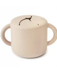 The mushie Snack Cup in Shifting Sand is a silicone cup with two handles and a flexible, slit lid for easy snack access and spill prevention, displayed on a white background.