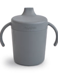 The Mushie Trainer Sippy Cup is gray, non-toxic, and features easy-grip handles with a spout lid. "mushie" is embossed on the front.
