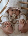 A baby in a cream hooded outfit with pom-pom trim sits on a soft surface, holding their feet and sucking on a FRIGG Natural Rubber Pacifier from the 4-Pack, looking off camera.