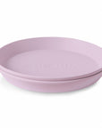 A light purple, round mushie Round Dinnerware Plate with raised edges and "mushie" embossed in the center, made from non-toxic plastic, is shown on a white background.