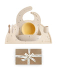The mushie Stage 1 Bees Feeding Kit—a beige silicone set with bib, bowl, cup, spoon, and fork, all adorned with small floral prints—sits on a matching placemat above a brown gift box tied with white ribbon. Ideal baby mealtime essentials.