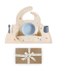 The mushie Stage 1 Dinos Feeding Kit features a beige silicone bib, bowl, cup, spoon, and fork with colorful dinosaur designs on a matching placemat. Below sits a brown gift box with a white “mushie” ribbon.