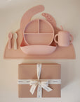 The mushie Stage 2 Blush Feeding Kit includes a divided plate, bowl, training cup with straw, silicone utensils, bib, and placemat in pink, all displayed above a kraft gift box tied with a branded ribbon bow.