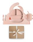 The mushie Stage 2 Blush Feeding Kit features a pink silicone bib, divided plate, bowl, cup with handles, training cup with straw, spoon, and fork on a matching placemat above a brown gift box tied with a white ribbon.
