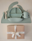 The mushie Stage 2 Cambridge Blue Feeding Kit includes silicone baby utensils, bib, divided plate, bowl, and training cup with straw on a matching placemat, all displayed above a gift-wrapped box with a white ribbon.