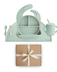 The mushie Stage 2 Cambridge Blue Feeding Kit includes silicone baby utensils, a bib, divided plate, training cup with straw, two spoons, and a matching placemat—all displayed above a brown gift box tied with a white ribbon.