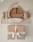 The mushie Stage 2 Natural/Shifting Sand Feeding Kit—featuring a divided plate, utensils, training cup, and straw—is neatly displayed above a gift box with a white ribbon against a light background.