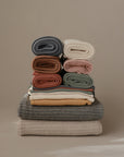 A stack of Mushie Ribbed Baby Blankets in grey, beige, cream, pink, and sage offers comfort and breathability; the soft blankets are folded and rolled, displayed against a taupe background.