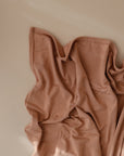 The mushie Stretchy Swaddle in a soft peach shade is draped with gentle folds and shadows on a smooth, light beige surface.