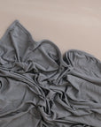 A mushie Stretchy Swaddle in light gray rests crumpled on a smooth beige surface, its breathable fabric forming soft, wavy folds and wrinkles.
