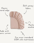 The mushie Stretchy Crib Sheet in blush pink is made from organic cotton, features a stretchy elastic band, breathable soft jersey fabric, is machine washable, and fits most standard USA crib mattresses.