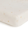 A close-up of the mushie Extra Soft Muslin Crib Sheet, featuring a white background with small yellow sun illustrations. The breathable sheet is fitted snugly around the mattress corner.