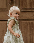 A young child with light blonde hair and a FRIGG Natural Rubber Pacifier 4-Pack by FRIGG stands before a weathered brown wooden door, wearing a light-colored sleeveless dress with a subtle pattern.