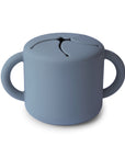 The mushie Snack Cup in Tradewinds is a gray silicone cup with two large side handles and a soft, flexible lid, designed for toddlers to easily access snacks while keeping them spill-proof.