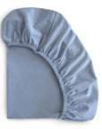 The mushie Stretchy Crib Sheet in light blue is partially folded with gathered elastic edges, displayed against a white background.