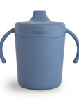 The Mushie Trainer Sippy Cup is a blue, non-toxic silicone sippy cup with easy-grip handles and a spout lid, featuring the "Mushie" brand on the front.