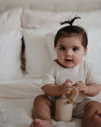 A dark-haired baby with a top knot sits on a white bed, holding a toy and wearing light clothes. Nearby is the FRIGG Natural Rubber Pacifier 4-Pack by FRIGG, offering comfort among fluffy pillows as the baby looks at the camera.