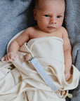 A baby wrapped in a Mushie Ribbed Baby Blanket lies on a soft gray surface, looking up with wide eyes. A pacifier with a wooden clip is attached to the breathable blanket near the baby's hand.