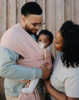 A man holds a baby in a pink wrap carrier, while a woman stands close, smiling. The baby, using the FRIGG Baby's First Pacifier Moonlight Sailing (Cream) 4-Pack by FRIGG, looks at the camera as they stand by a wooden wall.