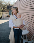 A woman smiles outside by a pink wall, holding a baby in a beige wrap carrier. The baby uses the Mushie FRIGG Rope Natural Rubber Pacifier 2-Pack. Nearby are outdoor chairs, a table, and trees in the background.