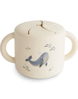 The mushie Snack Cup in Whales features dual handles, a spill-proof flexible lid, and a charming whale print—ideal for toddlers to snack easily on the go.