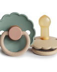 Two FRIGG Daisy Natural Rubber Pacifiers are displayed side by side on a white background; one is green with a beige handle, the other beige with a yellow latex nipple. The brand name FRIGG appears on both.