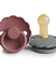 The FRIGG Daisy Natural Rubber Pacifier 2-Pack features two daisy-shaped pacifiers with scalloped edges, each marked with the FRIGG brand. One is mauve with a round handle, and the other is gray with a beige rubber nipple.