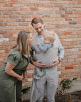 A smiling man carries a toddler in the mushie Baby Wrap against his chest, while a woman in green leans in and smiles at the child. Together, they share a joyful moment before a brick wall with small plants at their feet.