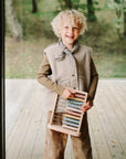 A smiling child with curly blond hair stands by a large window, holding the Mushie Wooden Abacus. The child is dressed in a beige vest, brown pants, and a checked scarf. Trees and greenery can be seen outside.