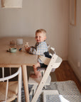 A young child sits smiling in a high chair at a wooden dining table, wearing a mushie Long Sleeve Bib. Bowls, cups, and utensils are on the table as sunlight streams through a nearby window.