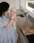A woman in a blue checkered dress holds a baby in a beige knit outfit and hat, who enjoys comfort with the FRIGG Butterfly Anatomical Silicone Pacifier 2-Pack (0-6 Months) by FRIGG, as they spend time together in the kitchen.
