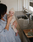 In a modern kitchen, a woman in a blue gingham dress offers her smiling baby—dressed in a beige knit outfit and hat—a mushie Fresh Food Feeder Freezer Tray for easy homemade baby food feeding and storage.