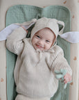 A smiling baby lies on a Mushie Changing Pad Liner, wearing a beige knitted outfit and a hat with bunny ears, holding a small toy.