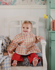 A toddler in red pants sits on a white chair, holding the Mushie Bath Rinse Cup. A patterned bib, wooden toy, and blue-and-white blanket complete the scene, with a daffodil in a pink jug atop a nearby mint-green cabinet.