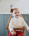 A smiling toddler with a red bow sits in a high chair, wearing the mushie Silicone Baby Bib featuring easy-to-clean material, small red patterns, and an adjustable neck strap. A blurred purple flower appears in the background.