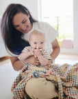 A smiling woman sits on the floor with a happy baby in her lap, wrapped in a mushie Organic Cotton Muslin Swaddle Blanket. Sunlight streams into the cozy indoor scene as the baby contentedly sucks on their fingers.