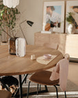 A round wooden dining table with minimalist decor, a pink bowl, and a cup—perfect for kids in the mushie Long Sleeve Bib. A light jacket hangs on a chair, art and plants decorate the sideboard, while a small dog naps under the table.