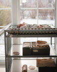 A baby rests on the mushie Extra Soft Muslin Changing Pad Cover atop a changing table by sunlit windows, with baskets of supplies below. Outside, snow blankets the ground and sunlight softly warms the cozy room.