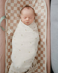 A newborn sleeps peacefully, wrapped in a mushie Organic Cotton Muslin Swaddle Blanket, lying on a beige and white checkered mat with soft pink and gray cushions on either side.