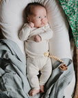 A baby in a cream outfit lies on a cushioned wicker bassinet, partially covered with the mushie Organic Cotton Muslin Swaddle Blanket in green floral; a wooden pacifier clip rests on the baby's waist.