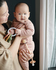 A woman smiles by a window, holding a baby dressed in pink. The baby looks at the camera while holding the mushie Western Teething Ring and a pacifier clip. Soft natural light brightens the scene.