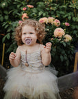 Toddler in a dress with FRIGG Daisy Natural Rubber Pacifier sits by blooming garden flowers.