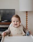 A smiling baby with light brown hair sits in a high chair, wearing a patterned bib from the mushie Stage 1 Dinos Feeding Kit and holding a gray teething toy. A record player, records, and a lamp are in the background.