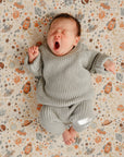 A baby in a cozy gray Mushie Chunky Knit Beanie and an organic cotton outfit lies yawning on a patterned blanket with space-themed designs, eyes closed and one fist slightly raised.