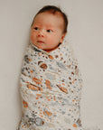 A baby lies on a soft white surface, wrapped in a mushie Organic Cotton Muslin Swaddle Blanket featuring orange, gray, and brown space-themed patterns, gazing slightly to the side.