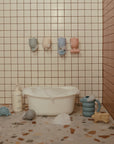 A small white bathtub with bubbles sits on a terrazzo floor in a tiled bathroom. Nearby are children’s bath toys, including the mushie Pour and Play Bath Set, a soap bottle, and four pastel cups hanging on the wall.
