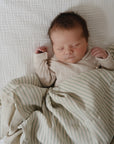 A newborn sleeps peacefully on a white textured blanket, wrapped in a mushie Organic Cotton Muslin Swaddle Blanket with green stripes. The baby, dressed in beige and with dark hair, looks serene.