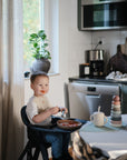 Smiling toddler with mushie Stacking Rings Toy in a cozy Danish Hygge kitchen.
