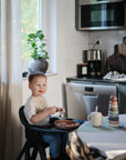 A young child sits in a kitchen, smiling with food on their face while wearing the mushie Silicone Baby Bib. They hold a spoon and bowl, with a cup and stacking toy on the table as sunlight streams through the window.